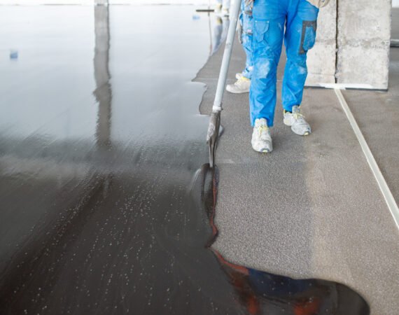 High angle of crop unrecognizable male workers in uniform and sneakers pouring and leveling liquid concrete on floor during renovation works at construction site