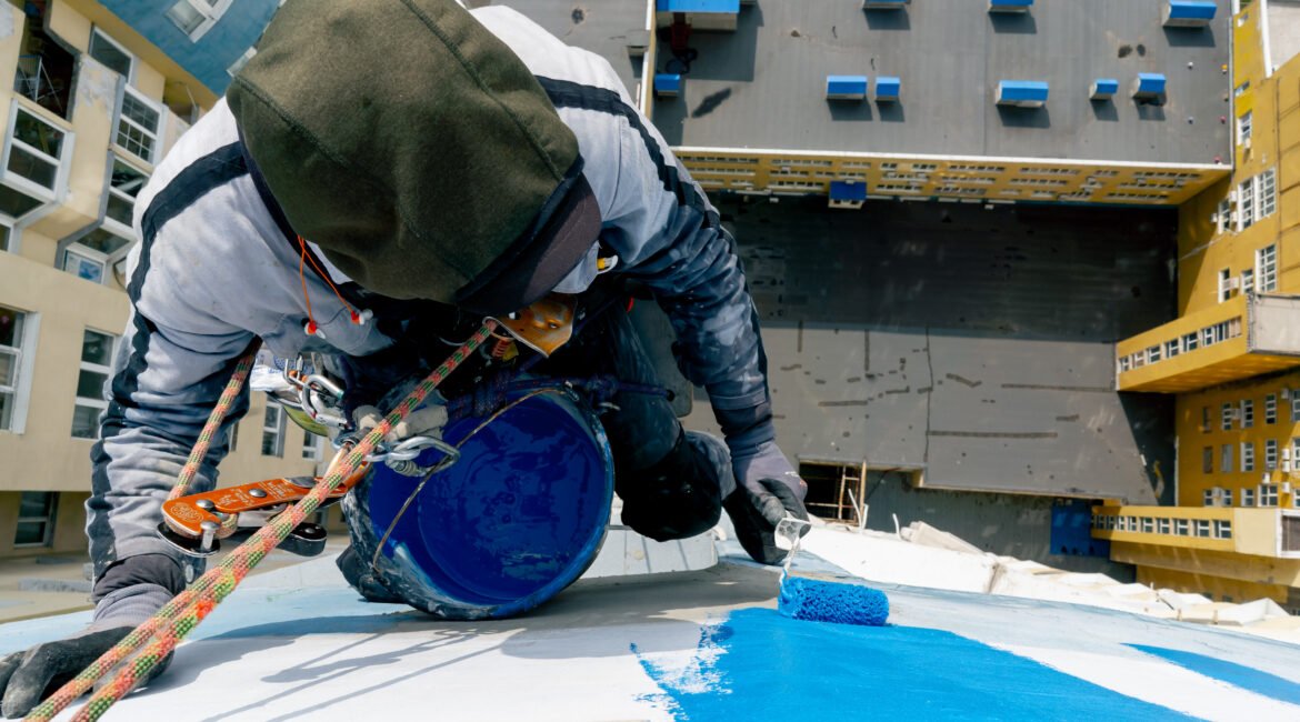 on the roof of a high-rise building against the background of multi-colored bright houses, a painter suspended on a safety net paints the wall blue